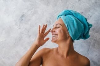 Smiling woman with a towel wrapped around her head applying facial cream.