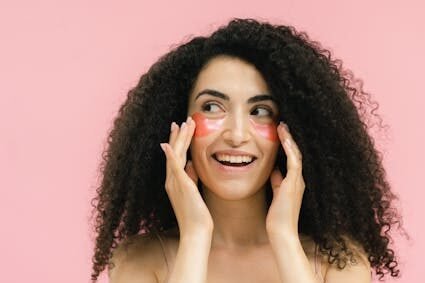 Happy woman with curly hair applying pink under eye patches on a pastel pink background.